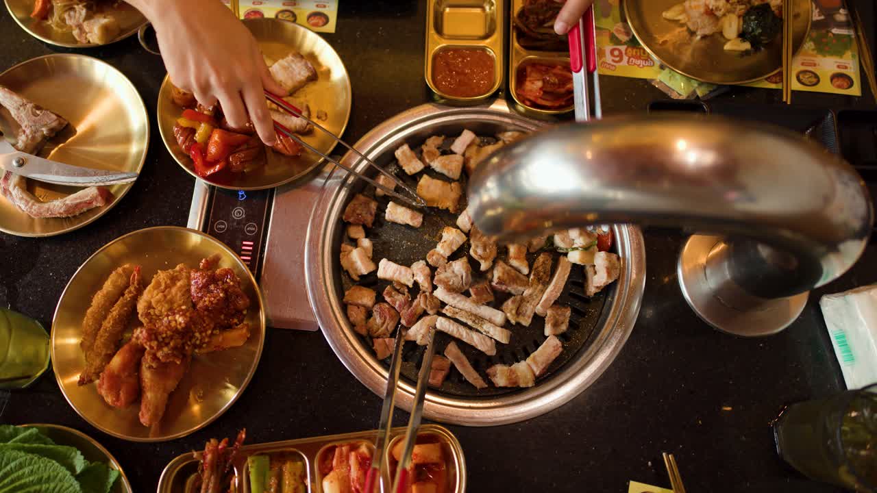 Hands use tongs to grill pork belly on tabletop Korean barbecue, overhead view, warm lighting