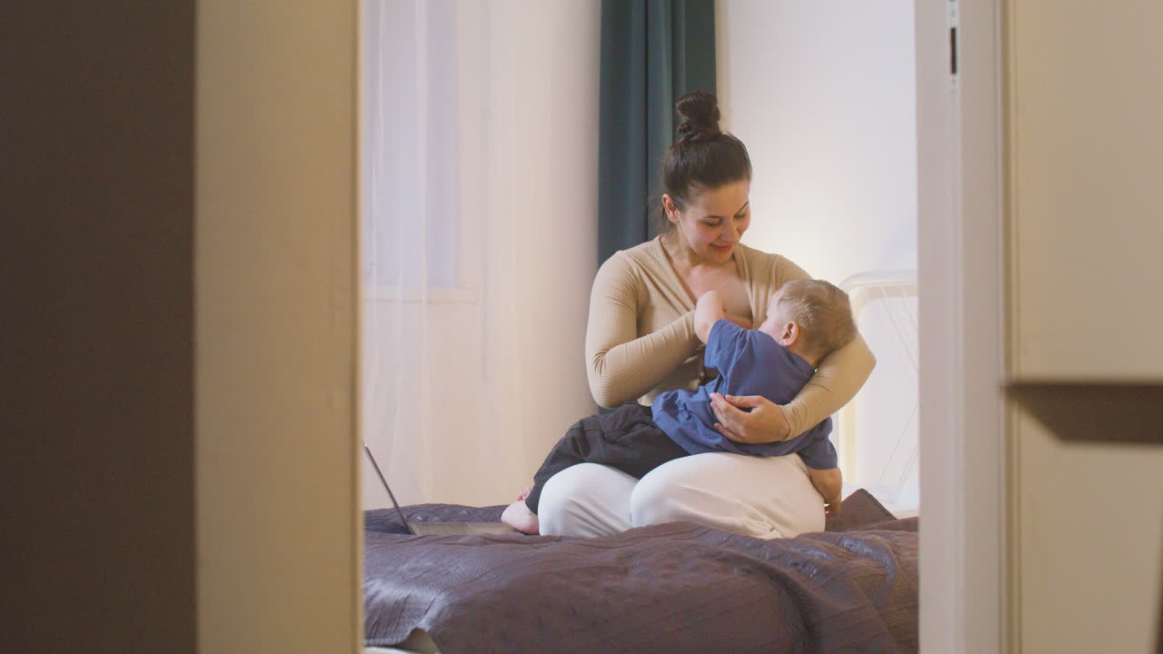 집에서 침대에 앉아있는 동안 랩톱 컴퓨터로 작업하고 아기를 모유 수유하는 젊은 어머니