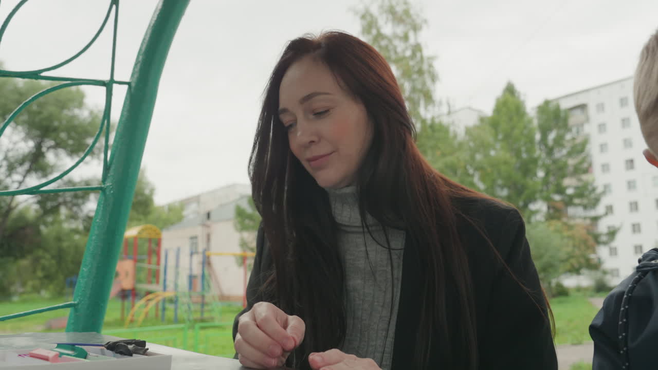 Donna e bambino che creano al tavolo del parco, sfondo di un parco giochi all'aperto, adulto che guida il bambino a preparare i materiali artistici per il ritorno a scuola con forme di argilla e pezzi colorati, mani concentrate, guida delicata.