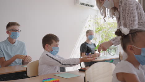 teacher children with face mask at school after lockdown disinfecting hands. school boy holding out his hands while sitting at desk in classroom as female teacher pouring some sanitize