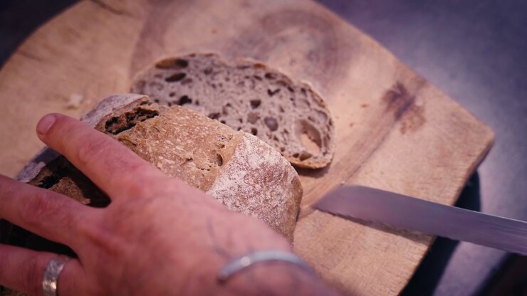French country bread being sliced by hand on a rustic wooden board. Close-up view with warm cinematic tones highlighting the artisanal bakery tradition in a domestic kitchen.