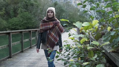 serene woman in outerwear walking on footbridge over tranquil lake