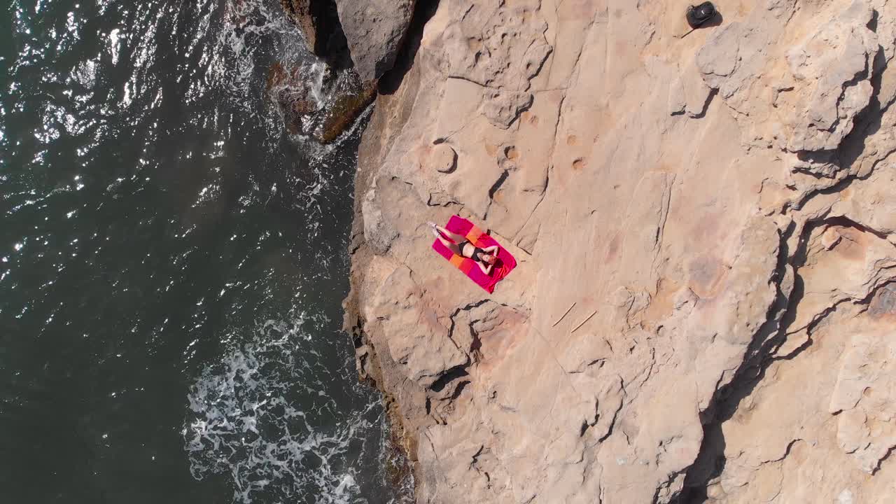 Aerial, top down, drone shot of a woman lying on a towel, while waves hit the rocky coast of Mallorca, sunny day, in Spain