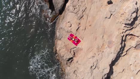 aerial, top down, drone shot of a woman lying on a towel, while waves hit the rocky coast of mallorca, sunny day, in spain