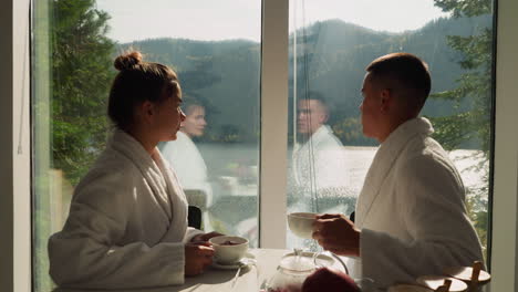 couple drinks tea looking at window view. newlyweds in terry bathrobes enjoy tea-time admiring nature around lakeside hotel. perfect weekend morning