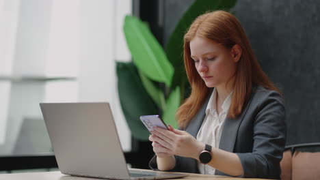 pretty redhead woman is working in office of big modern company using private smartphone on workplace