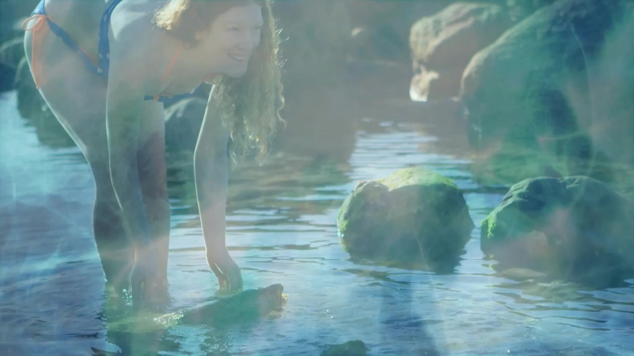 Mulher caminhando em piscina natural, coletando e explorando a água, com ondulações e um brilho azul-esverdeado realçando a experiência de viagem.