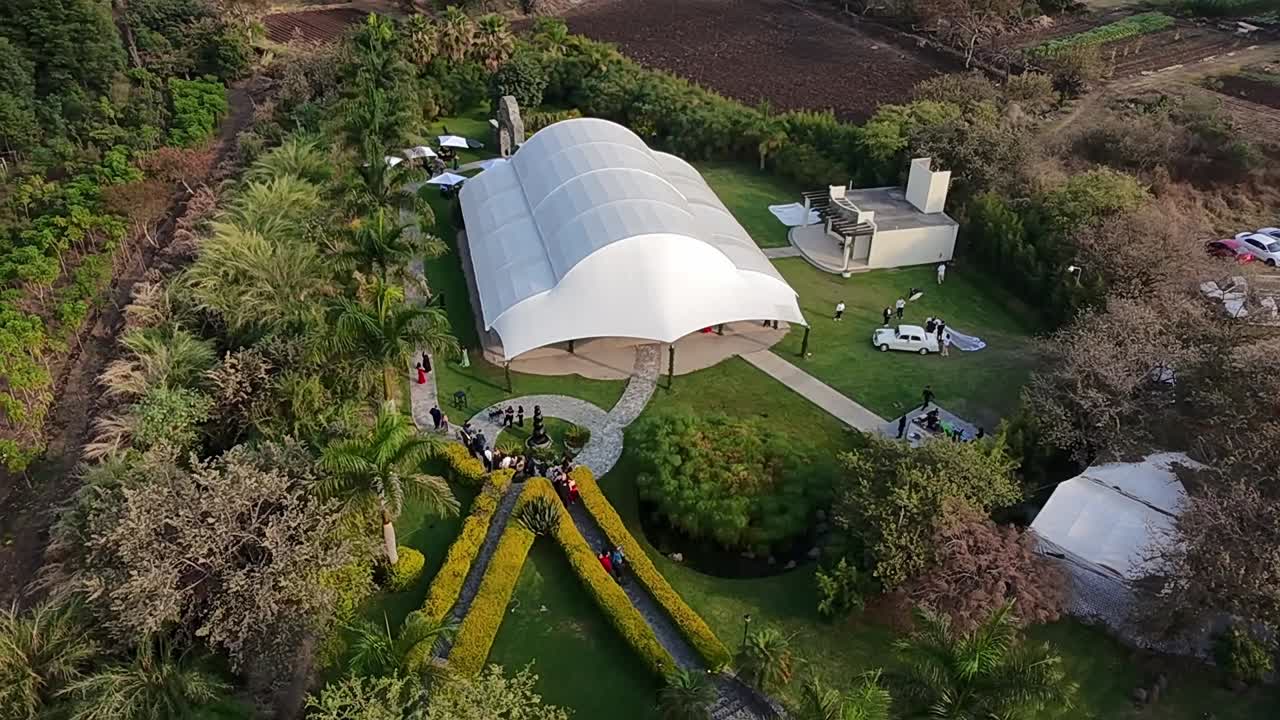 erial slow-motion orbit over Jardín Villa Cortés, Morelos, showing guests at a wedding cocktail party.
