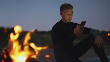 man sitting on surfboard by camp fire on beach using mobile phone as sun sets behind her