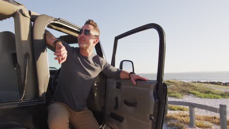 feliz hombre caucásico con gafas de sol sentado en el coche con la puerta abierta en un día soleado en la playa