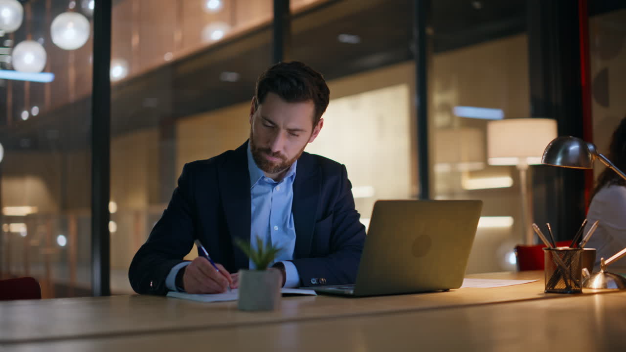 Primo piano di un manager che analizza i dati su un portatile seduto alla scrivania in ufficio di sera con un collega