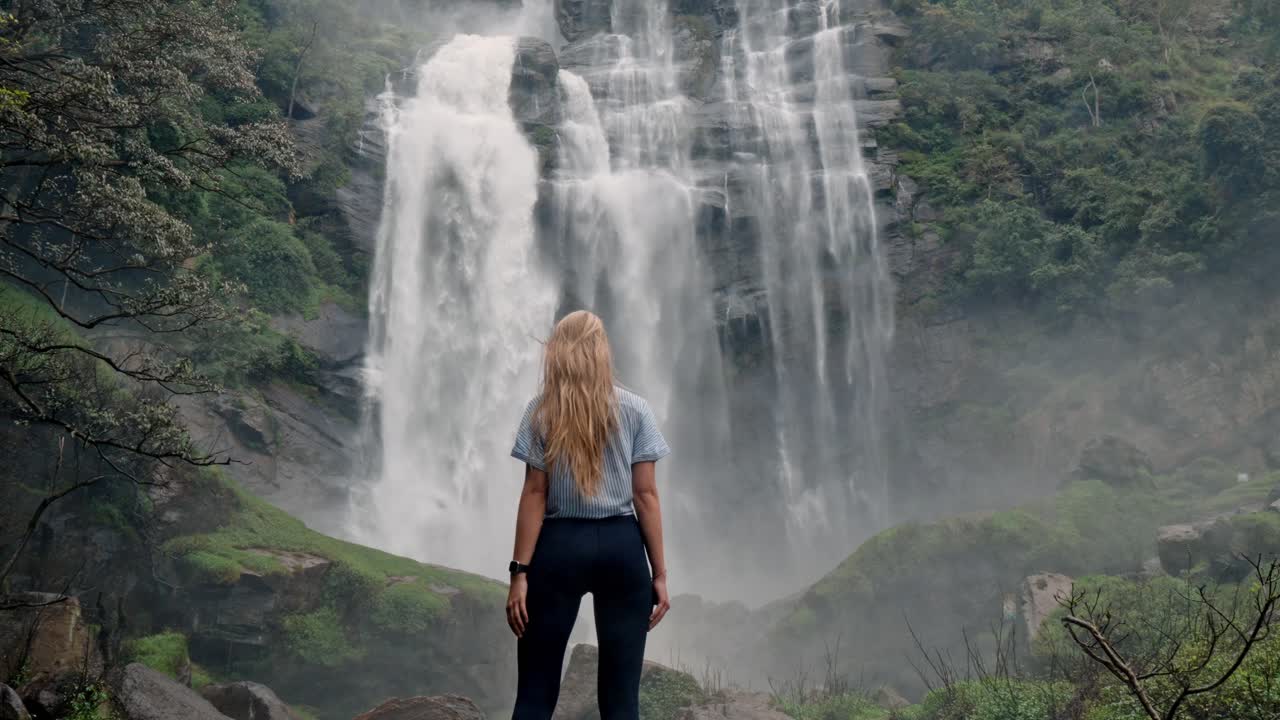 A woman stands in awe before the roaring Bomburu Ella Waterfalls, surrounded by mist, jungle, and rocky cliffs in the lush highlands of Nuwara Eliya, Sri Lanka.