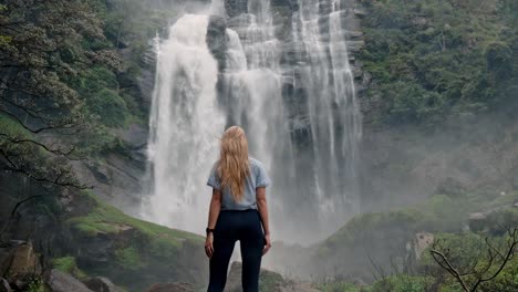 A woman stands in awe before the roaring Bomburu Ella Waterfalls, surrounded by mist, jungle, and rocky cliffs in the lush highlands of Nuwara Eliya, Sri Lanka.