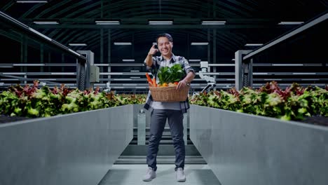 cuerpo lleno de granjero masculino asiático con canasta de verduras sonriendo y haciendo un gesto honesto en la granja inteligente
