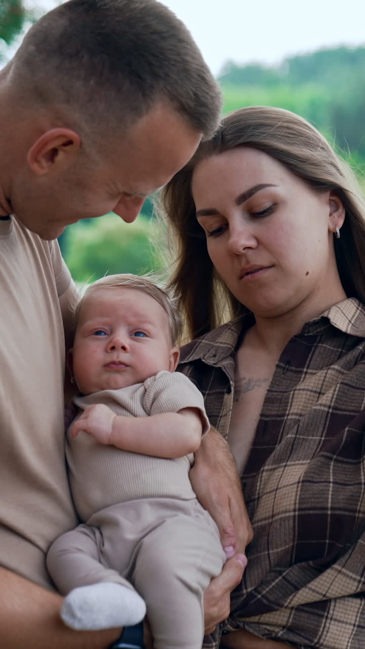 Caucasian couple with their infant baby standing outdoors. Mom and dad protecting their son from the wind. Mother kisses the baby on the head. Vertical video