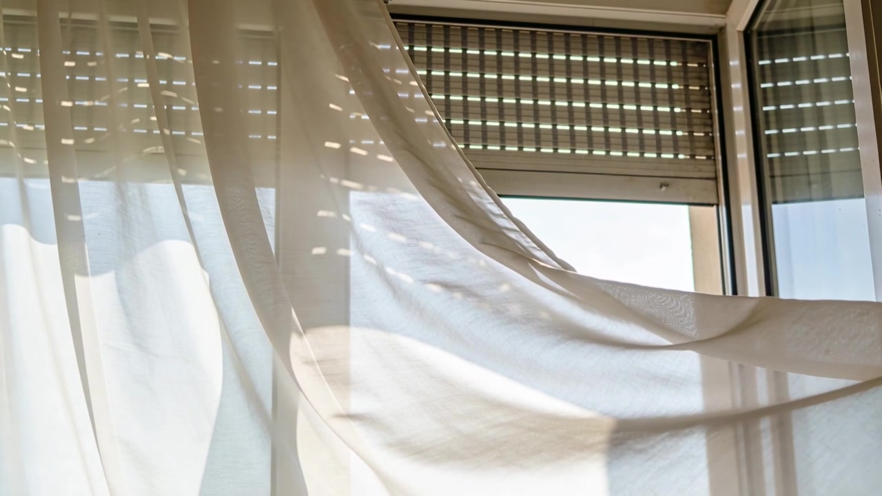Tenda trasparente che ondeggia, catturando una corrente d'aria da una finestra a battente aperta a casa, mostrando macchie di luce dalle persiane.