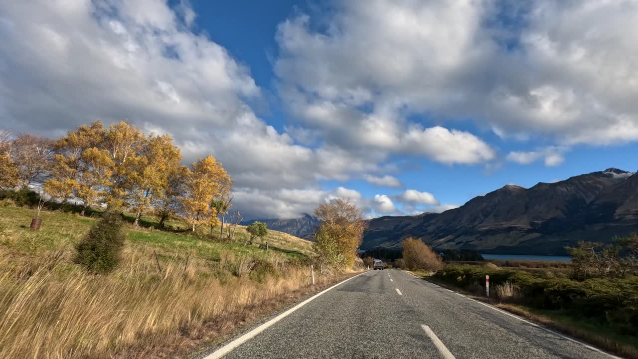Car travels rural road through autumn landscape, mountains, and lake under partly cloudy daylight sky