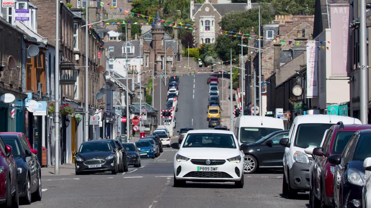 White car drives toward camera on busy, sunlit Scottish street with shops and parked vehicles