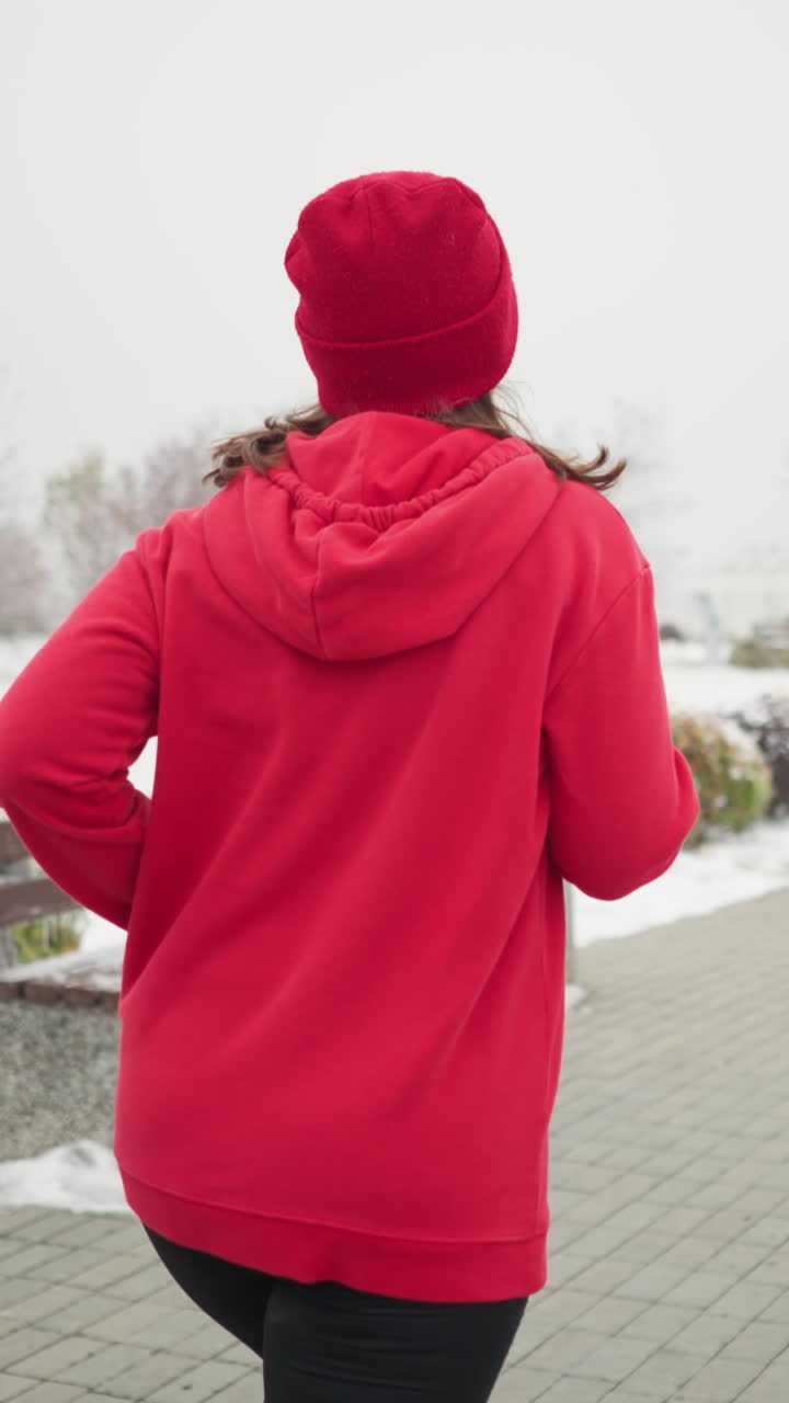 back view of woman jogging in red beanie and jacket along snowy park pathway with iron railing benches nearby serene foggy winter atmosphere frosty trees distant snow-dusted building