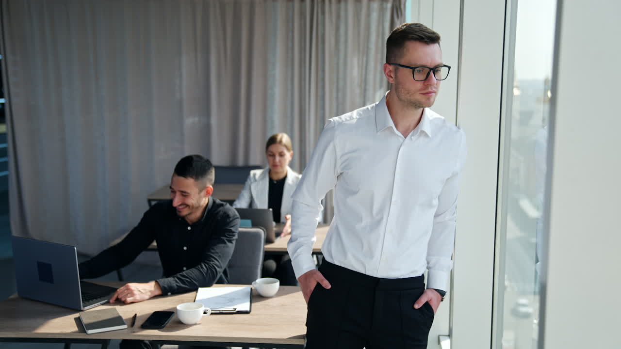 Calm peaceful pensive man standing at the window thinking over something. Colleagues working at laptops at backdrop.