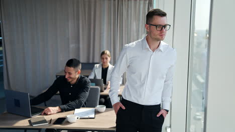 calm peaceful pensive man standing at the window thinking over something. colleagues working at laptops at backdrop.