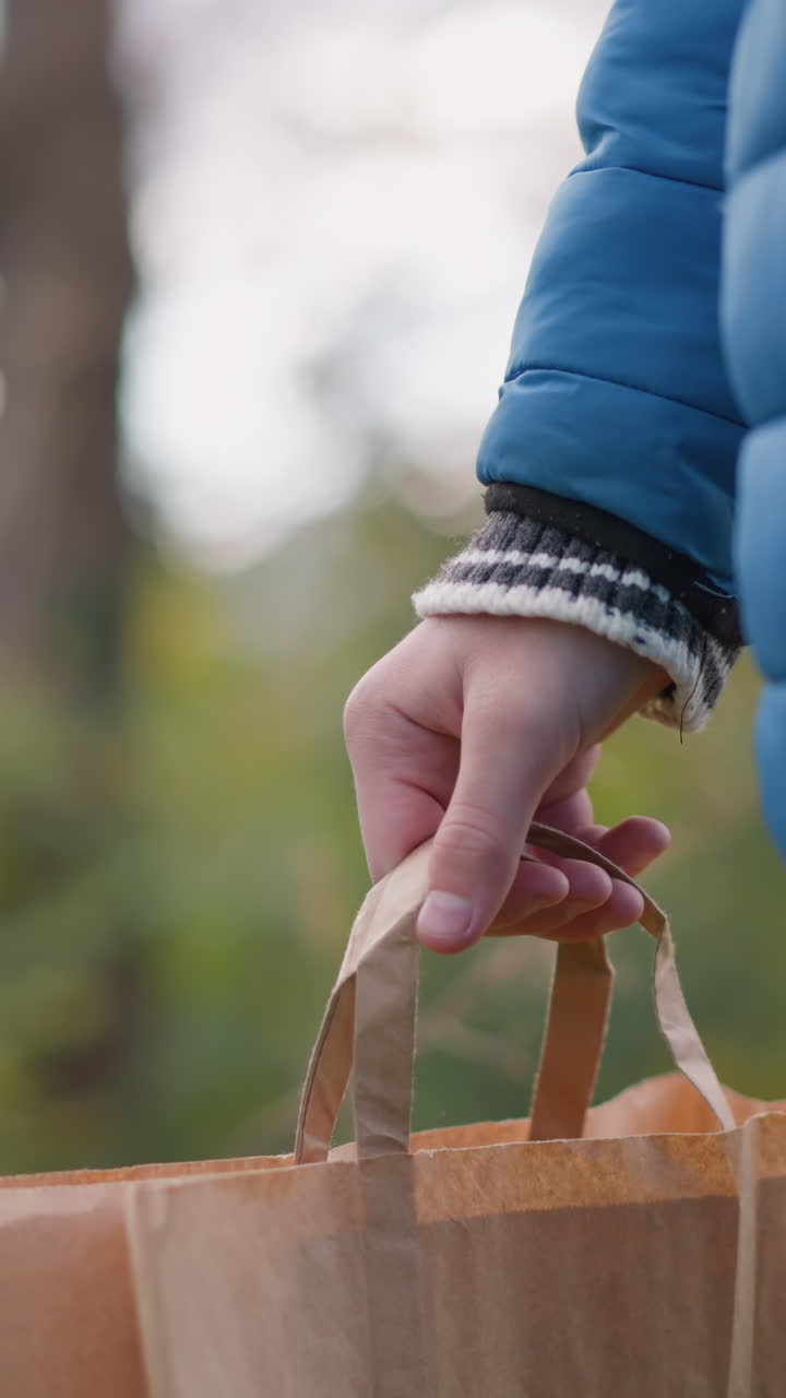vista parcial de cerca de una persona con chaqueta azul caminando al aire libre sosteniendo una bolsa de papel marrón, balanceando el brazo casualmente, rodeada de vegetación vibrante y elementos naturales