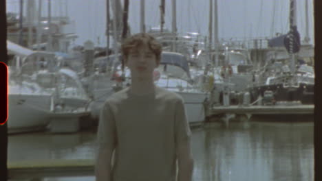 filmed on super 8 film - a young man standing on a pier in a marina, with sailboats and yachts moored in the background, filmed in a retro style