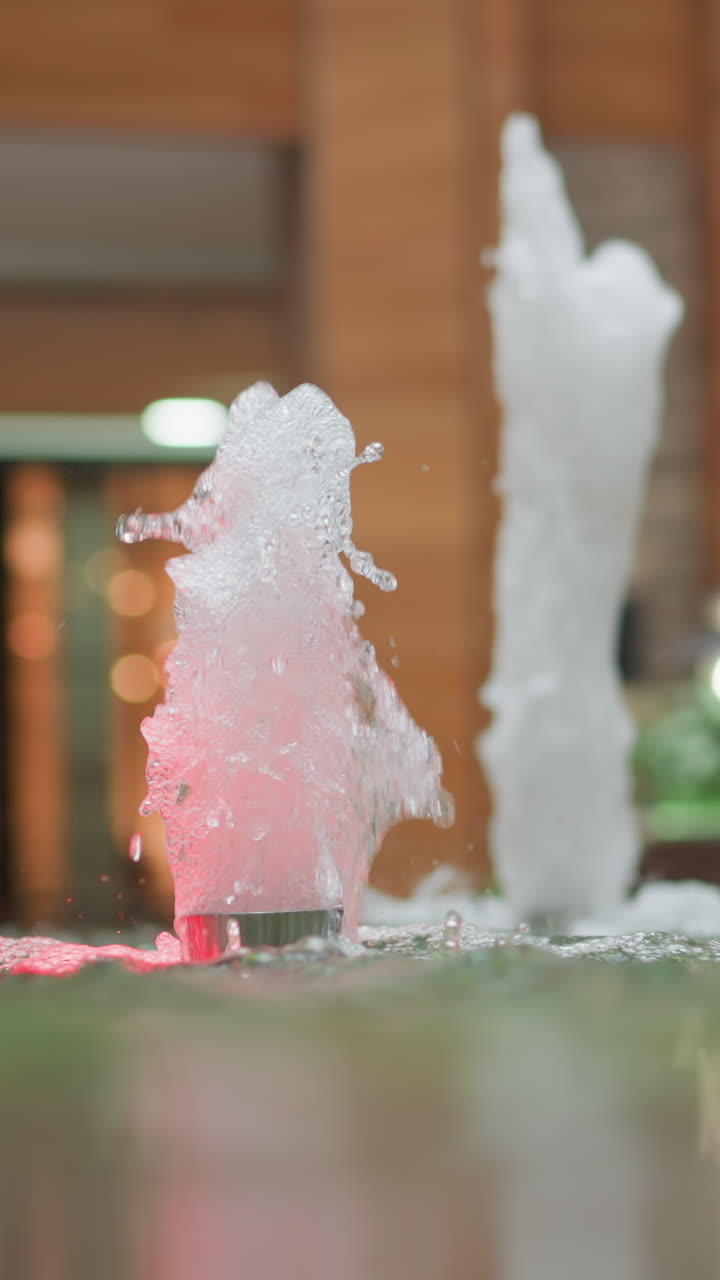 타일로 된 기둥, 에스컬레이터, 울창한 초록색으로 둘러싸인 현대 쇼핑몰 환경에서 다채로운 불빛으로 조명된 활기찬 하천으로 된 실내 물 분수