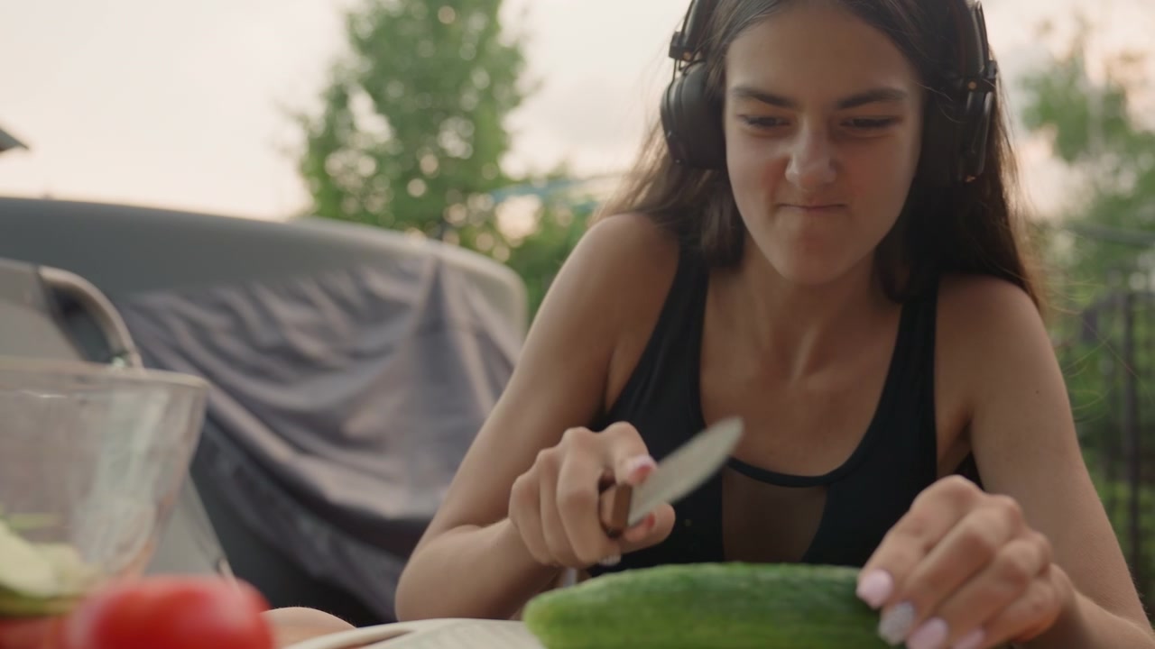 Giovane donna con le cuffie prepara verdure all'aperto, affettando un cetriolo su un tagliere con prodotti freschi visibili. La donna è seduta a un tavolo esterno vicino alla vegetazione e a una luce naturale soffusa