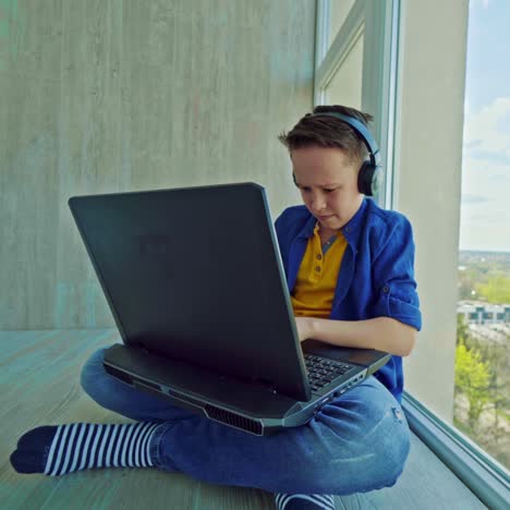 teenager plays games on a modern gadget at home. boy with earphones sitting on the floor near the window and becomes nervous while playing video games on a laptop.