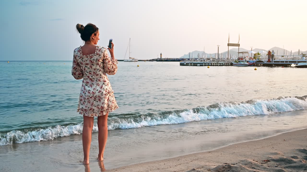 Blonde woman shooting on her smartphone on the beach on the Mediterranean sea coast in Cannes, France at sunset