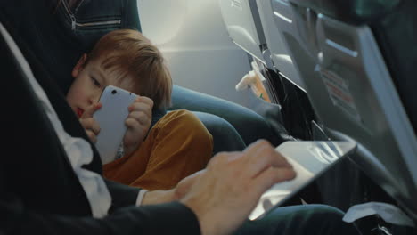 Boy-using-smart-phone-and-father-with-tablet-in-plane