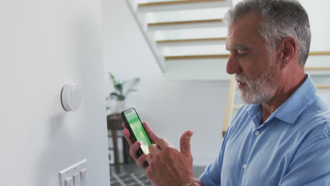 un anciano hispano usa una aplicación en su teléfono para controlar el termostato digital de calefacción central en casa.