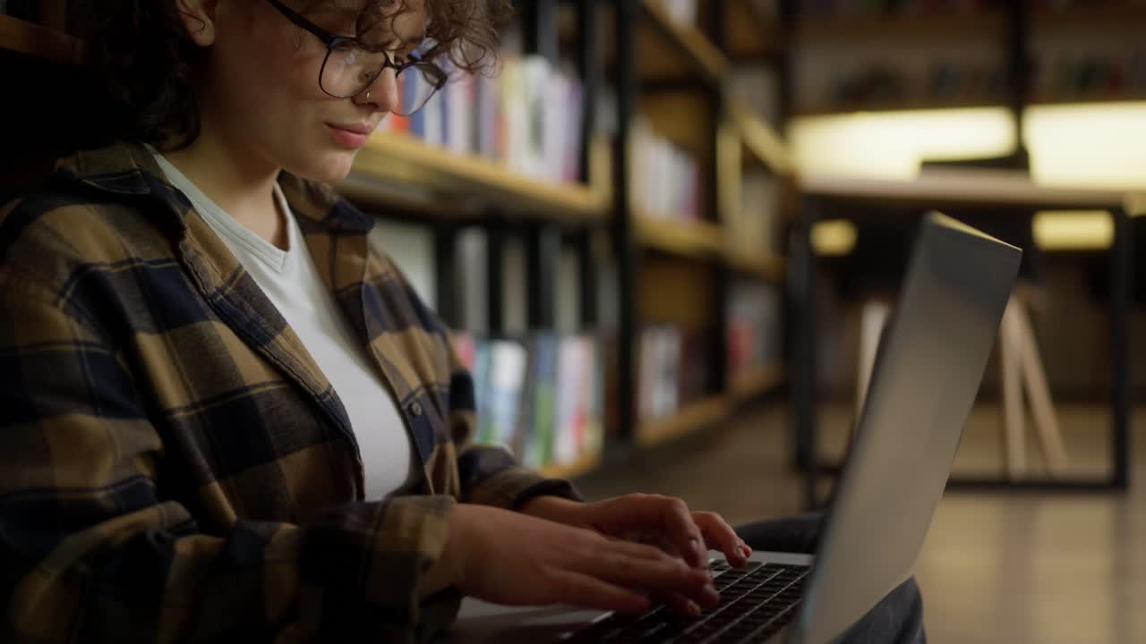 cerca de una estudiante con gafas con cabello rizado en una camisa a cuadros hace notas en una computadora portátil gris mirando las estanterías con libros en la universidad