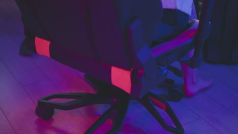 lower leg view of person wearing fishnet pantyhose stepping across neon lit floor toward rolling chair at gaming desk with monitors and lights under pink purple ambient glow before sitting down