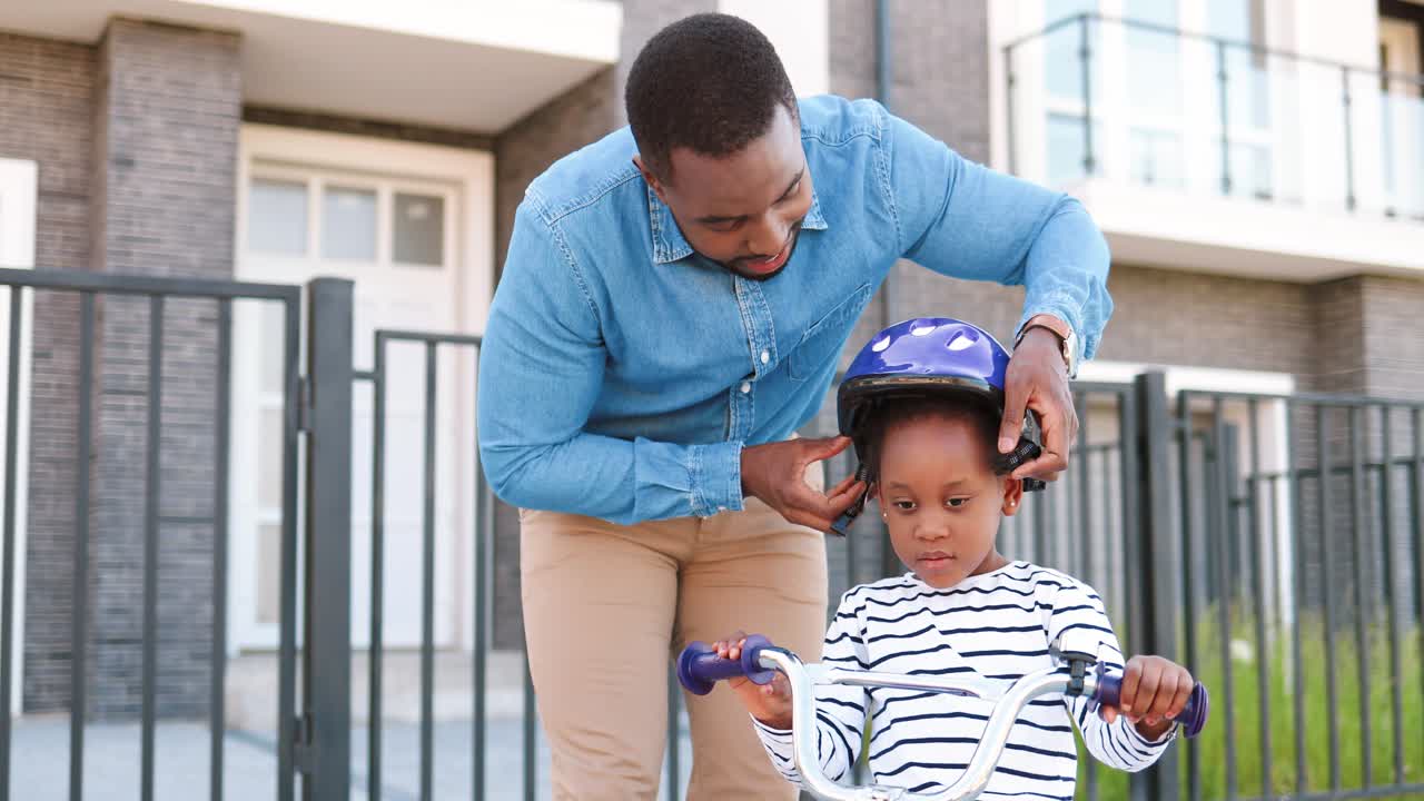 African American father putting helmet on small pretty girl and teaching ride on bike at street