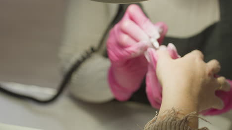 client hand with gold ring receiving manicure as technician in pink gloves uses electric nail drill. technician appears blurred in background while filing tool focuses on precision nail care routine