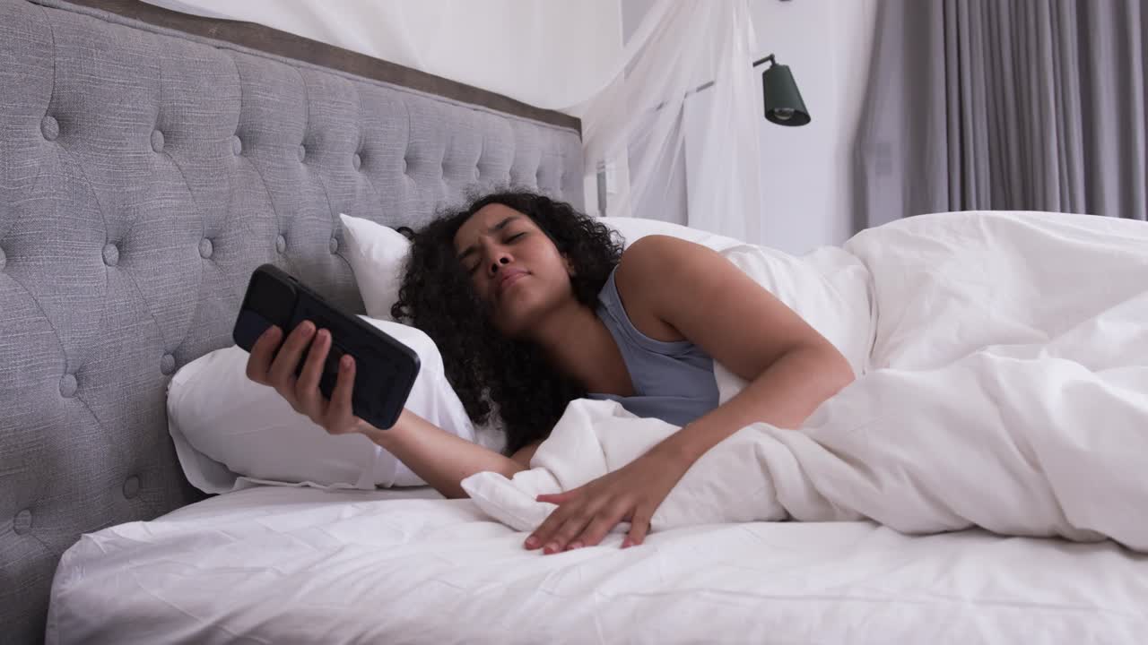 Afroamerikansk kvinde, der vågner, rækker ud efter telefonen og justerer dynen i sengen iført en blå tanktop.