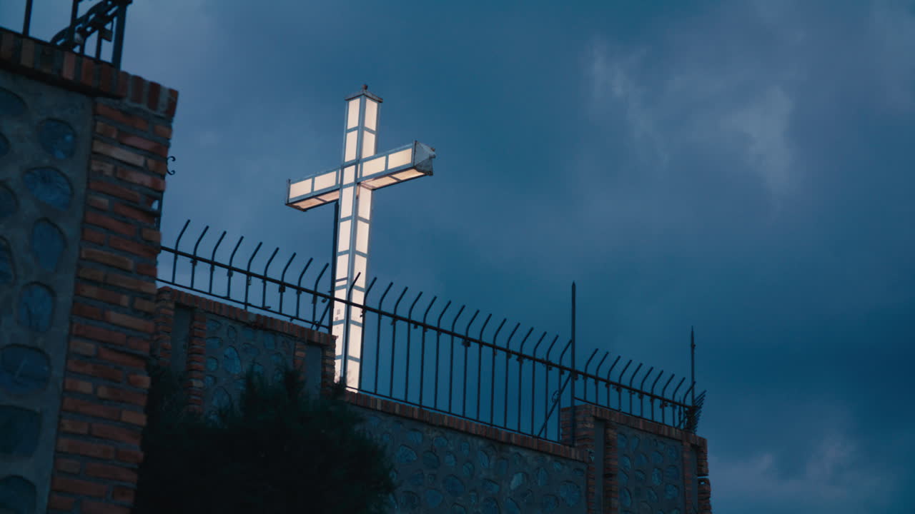 Croce luminosa e illuminata che si erge sopra un muro di pietra e una recinzione di ferro, brillando intensamente contro un cielo tempestoso blu scuro, creando un forte contrasto visivo e un'immagine simbolica di luce nell'oscurità.