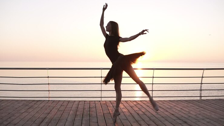 Young stunning ballerina on a wooden seafront doing ballet pas. Morning. Girl wearing dark tutu. Slow motion