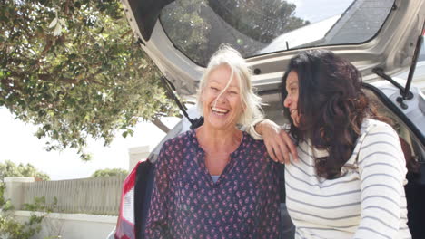 Two-happy-diverse-senior-women-sitting-in-open-trunk-and-embracing-on-sunny-day,-slow-motion