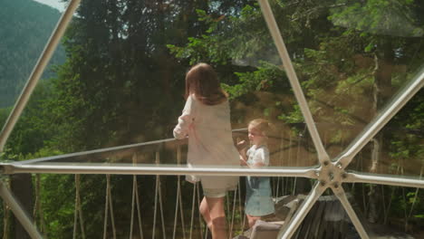 mother and daughter stand taking photos on glass balcony. little girl holds camera spending time against dense forest. happy family photograph against glamping park