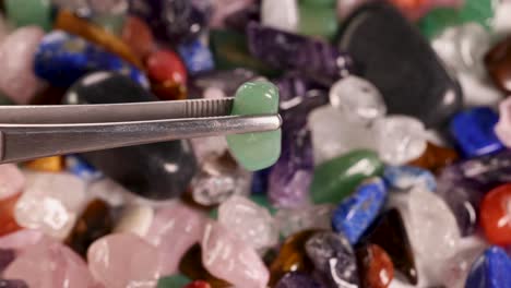 A green aventurine stone is carefully picked with tweezers from a vibrant assortment of chakra stones under bright lighting