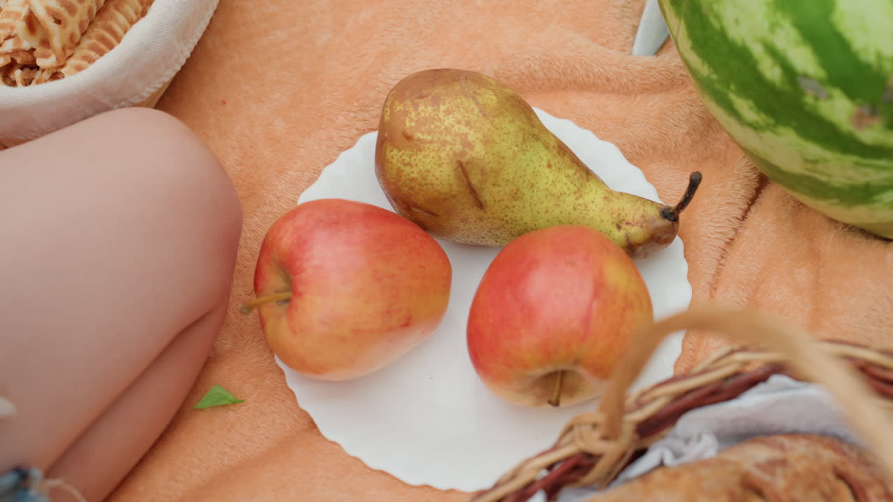 Primer plano de un picnic con peras y manzanas sobre una toalla naranja junto a las piernas de una persona, borde de pantalones cortos de mezclilla, cesta de mimbre y sandía, ambiente tranquilo de ocio veraniego, fondo de hierba iluminada por el sol, snack natural y fresco