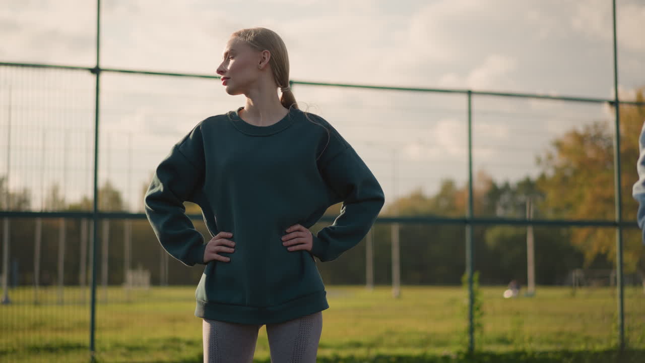 joven con suéter verde con la mano en la cintura, haciendo ejercicio girando la cabeza de izquierda a derecha, la luz del sol crea un resplandor dorado a su alrededor, con vista parcial de alguien a su lado en un entorno al aire libre