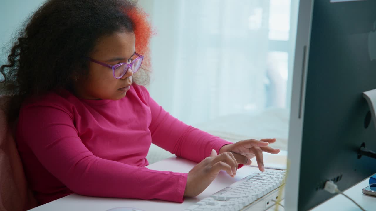 Young female student wearing glasses and colorful hair sitting at white desk, focusing intently while typing on computer keyboard during online learning session
