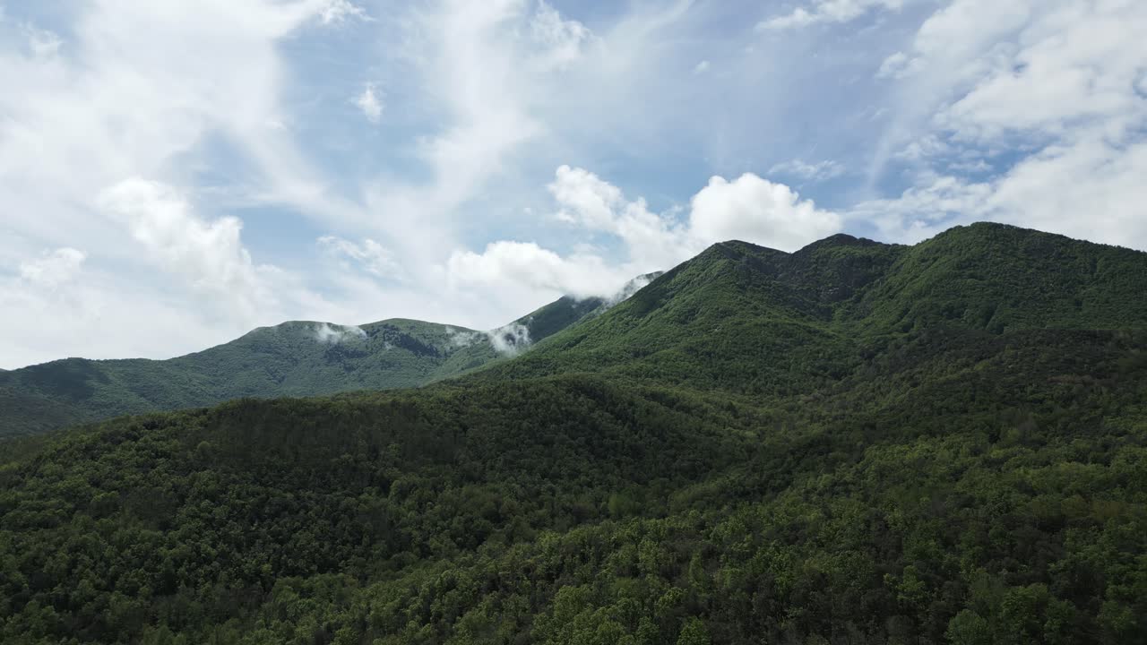 Verdant mountain range revealing pristine wilderness with white clouds drifting over luminous blue sky, showcasing breathtaking natural landscape through sweeping panoramic view