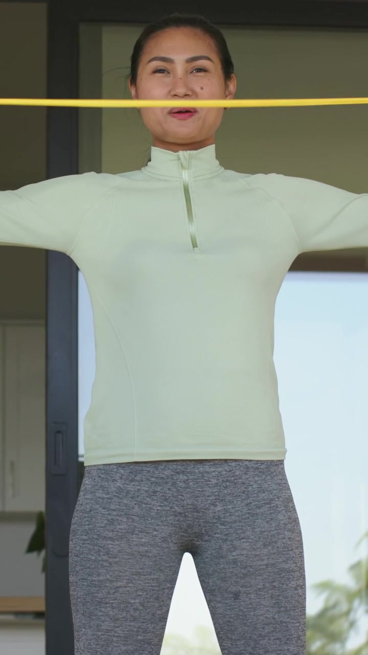 Vertical video: Pulling yellow band across chest, Asian woman toning shoulders at home in green top