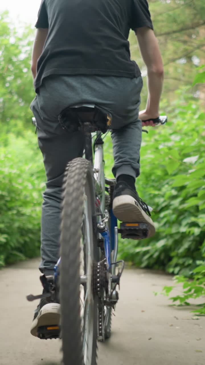 Back view of young boy wearing black top and grey trouser cycling along a paved path with white road markings, surrounded by vibrant greenery that forms a natural tunnel effect