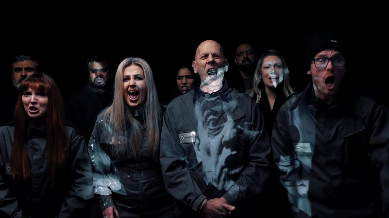 Persone che urlano verso uno schermo di propaganda si trovano in una drammatica penombra indossando uniformi abbinate. La scena di gruppo evoca la distopia orwelliana, i Due Minuti d'Odio, l'indottrinamento di massa e il controllo autoritario.